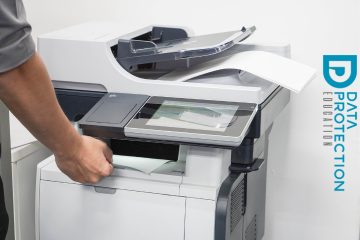 A hand taking some documents off of a large printer. The data protection education logo in blue going down from top to bottom to the right of the printer