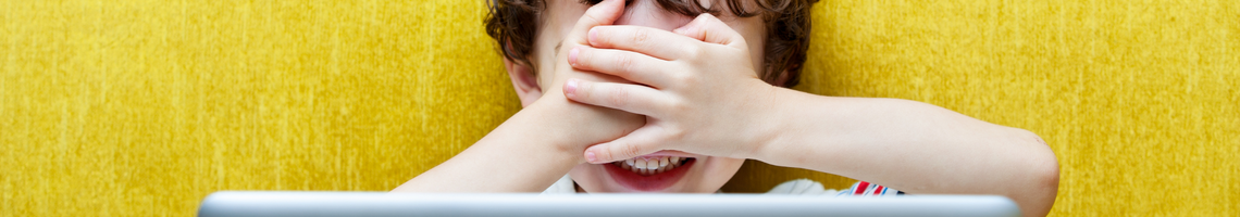 Child with their hands over their eyes in front of a computer laptop. Yellow background. White desk