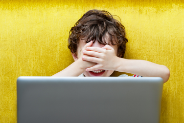 Yellow background. Young child in front of a laptop with their hands over their face